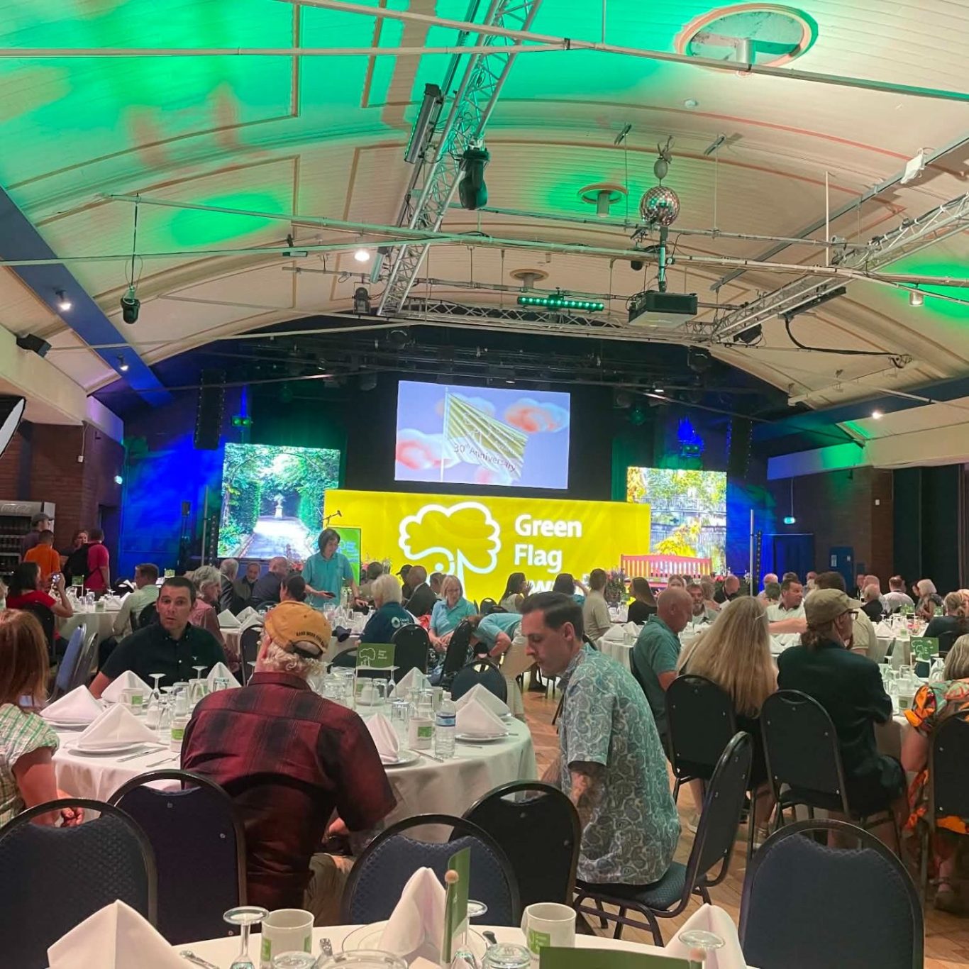 A banquet hall with green lighting, tables set for dining, and a presentation on stage.