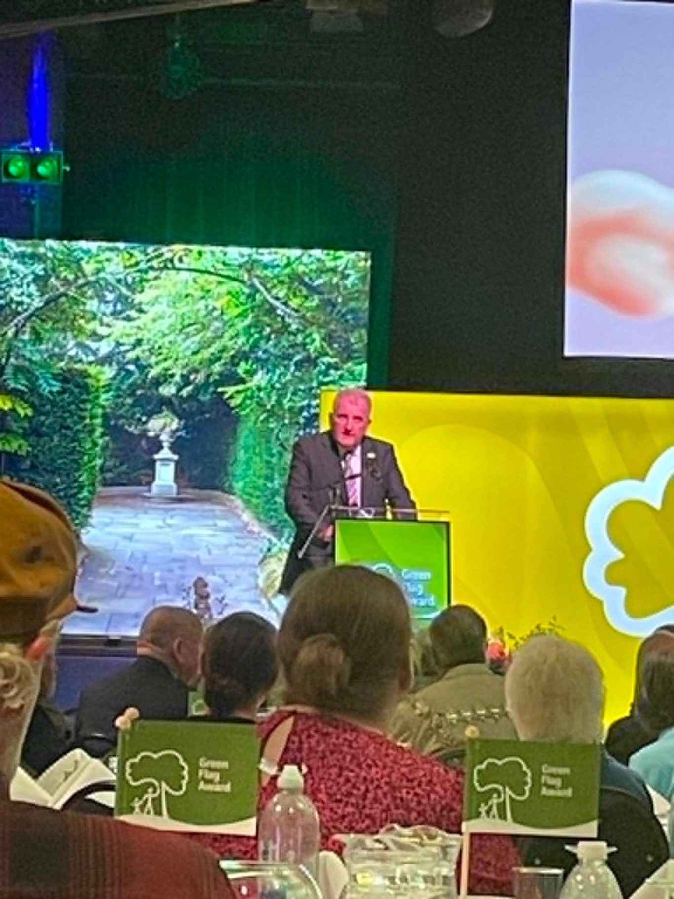 Event attendees seated at tables, with green tree-themed decorations and a stage backdrop.